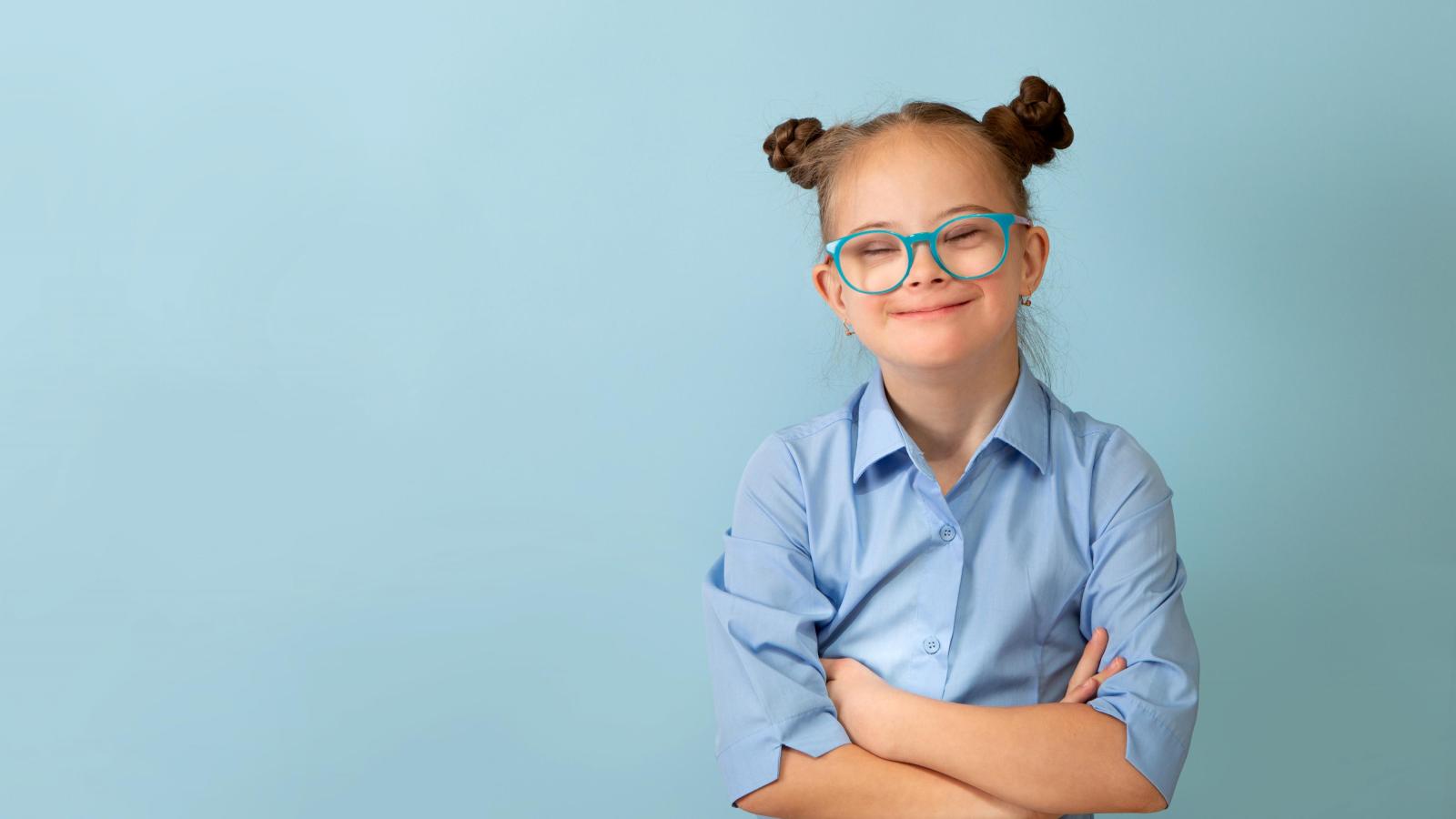 Mädchen mit Down-Syndrom steht lächelnd mit verschränkten Armen vor einem hellblauen Hintergrund