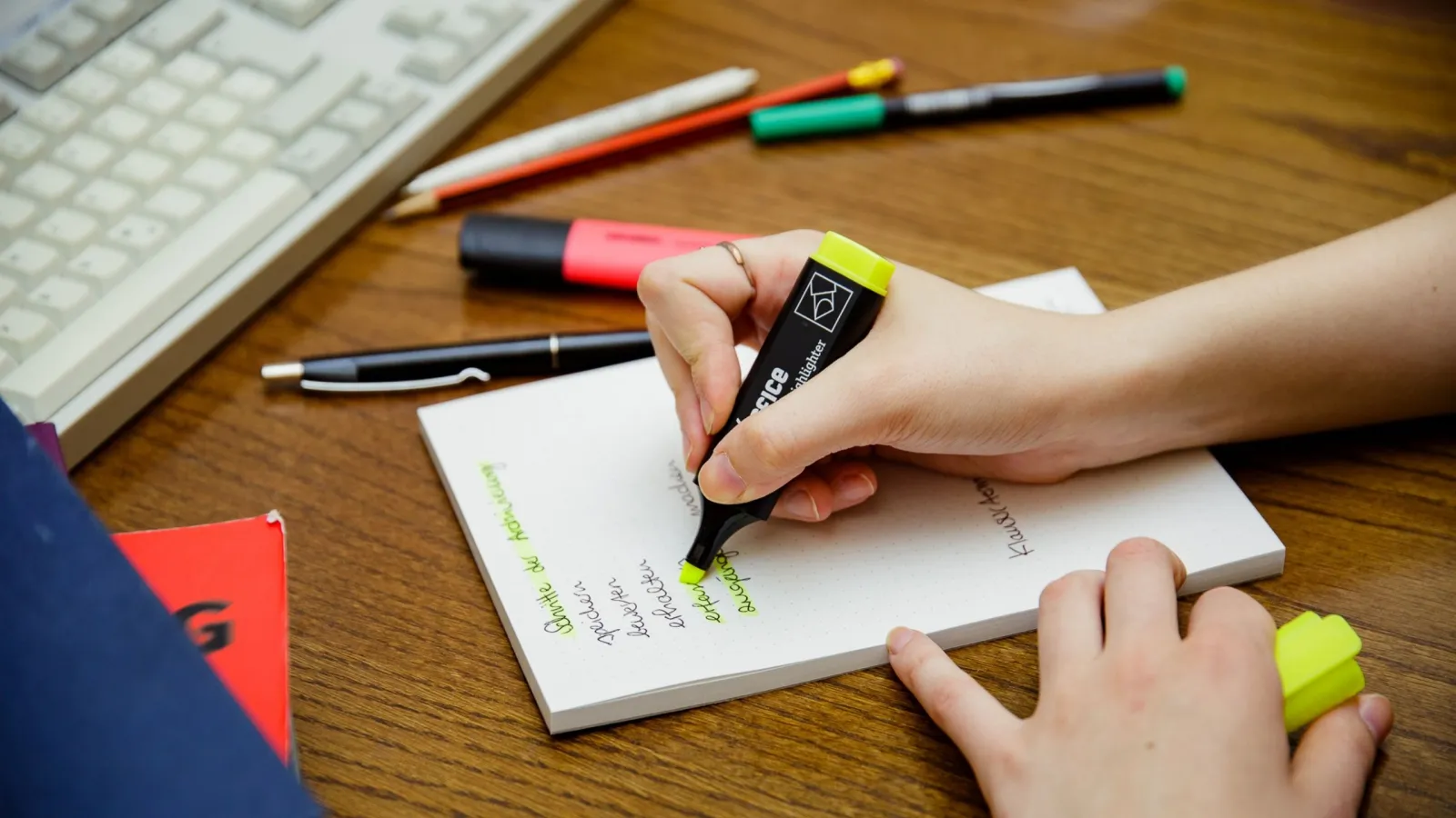 Studentin benutzt einen Textmarker, um in einer Liste zu markieren. Zu sehen ist die Tischoberfläche, ein Block, diverse Stifte, sowie die Hände. Im Hintergrund ist eine Tastatur zu erkennen.