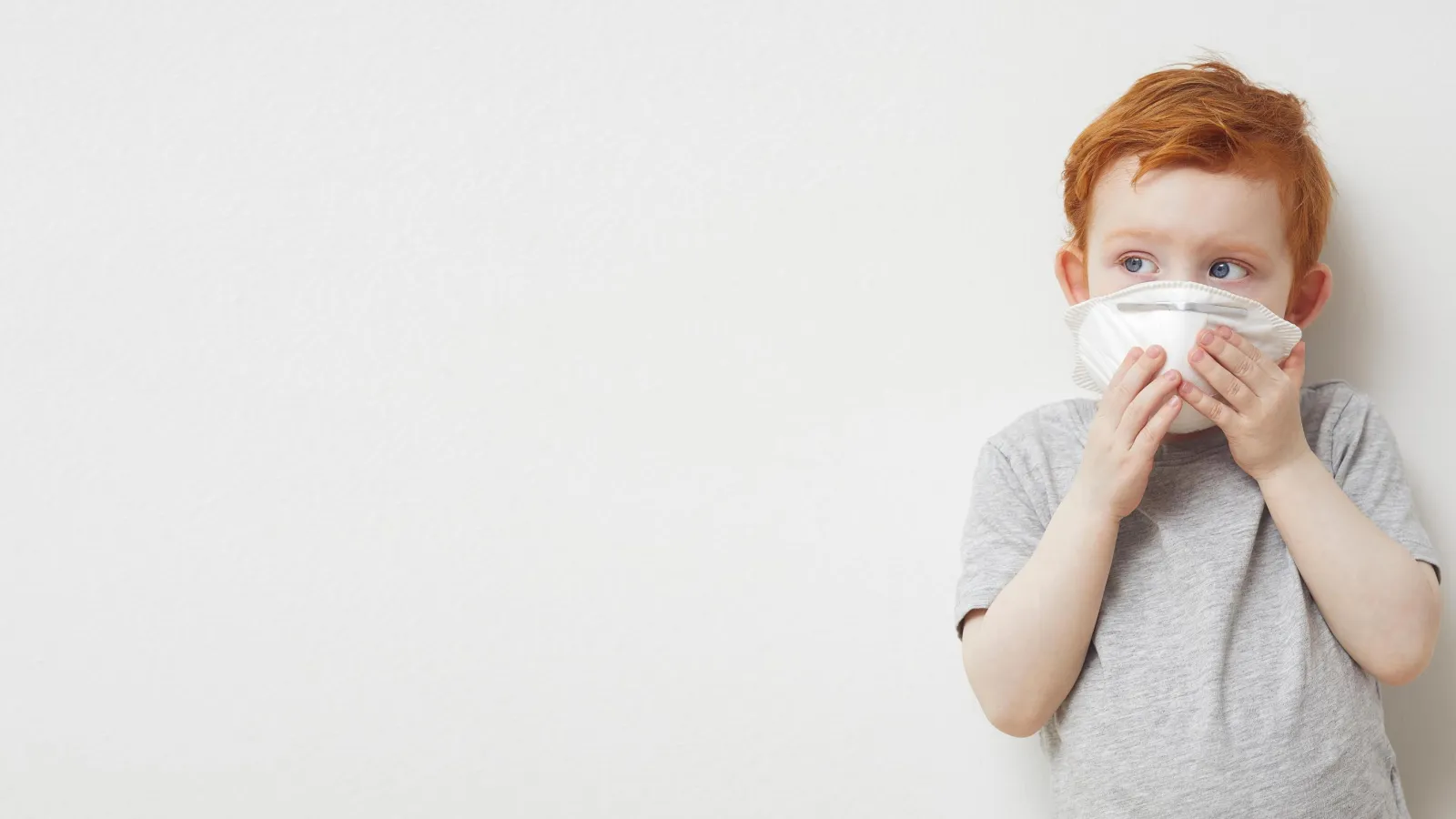 Junge mit Corona-Maske vor dem Gesicht, steht vor heller Wand