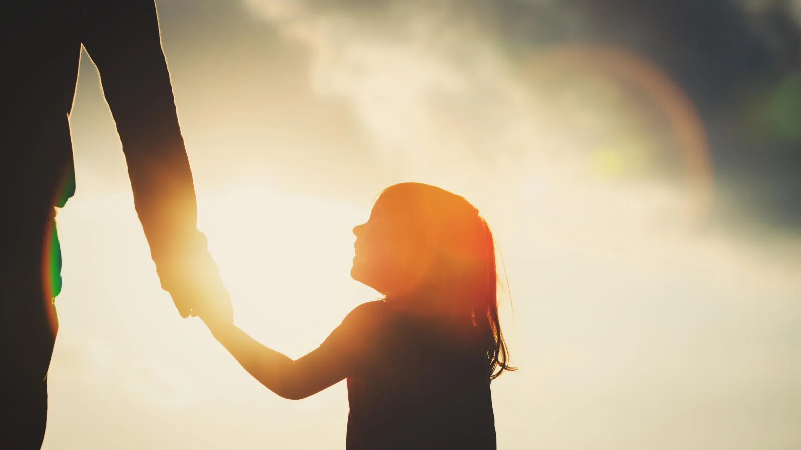 Silhouette eines Kindes, das an der Hand eines Erwachsenen läuft, im Hintergrund die Sonne