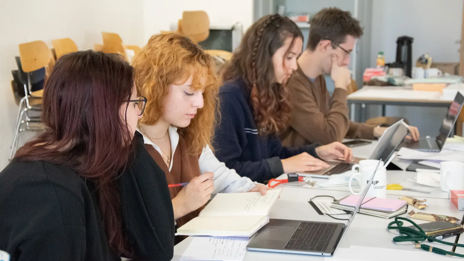 Vier Teilnehmende von der Seite sitzend am Tisch mit Notebook und Notizheften