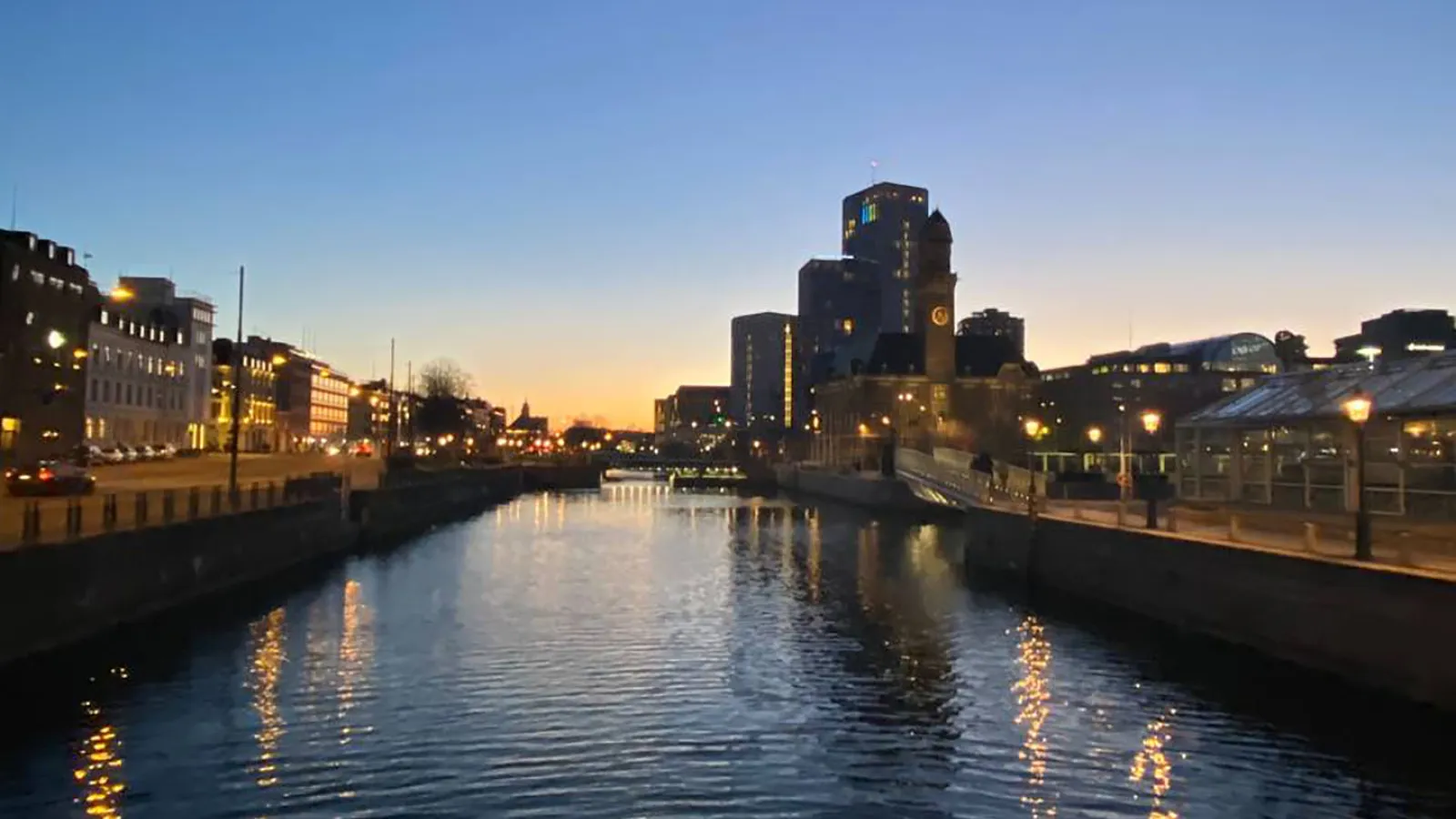 Ein Fluss beim Sonnenuntergang. Der Himmel ist wolkenlos. Die Lichter des Ufers spiegeln sich im Wasser.