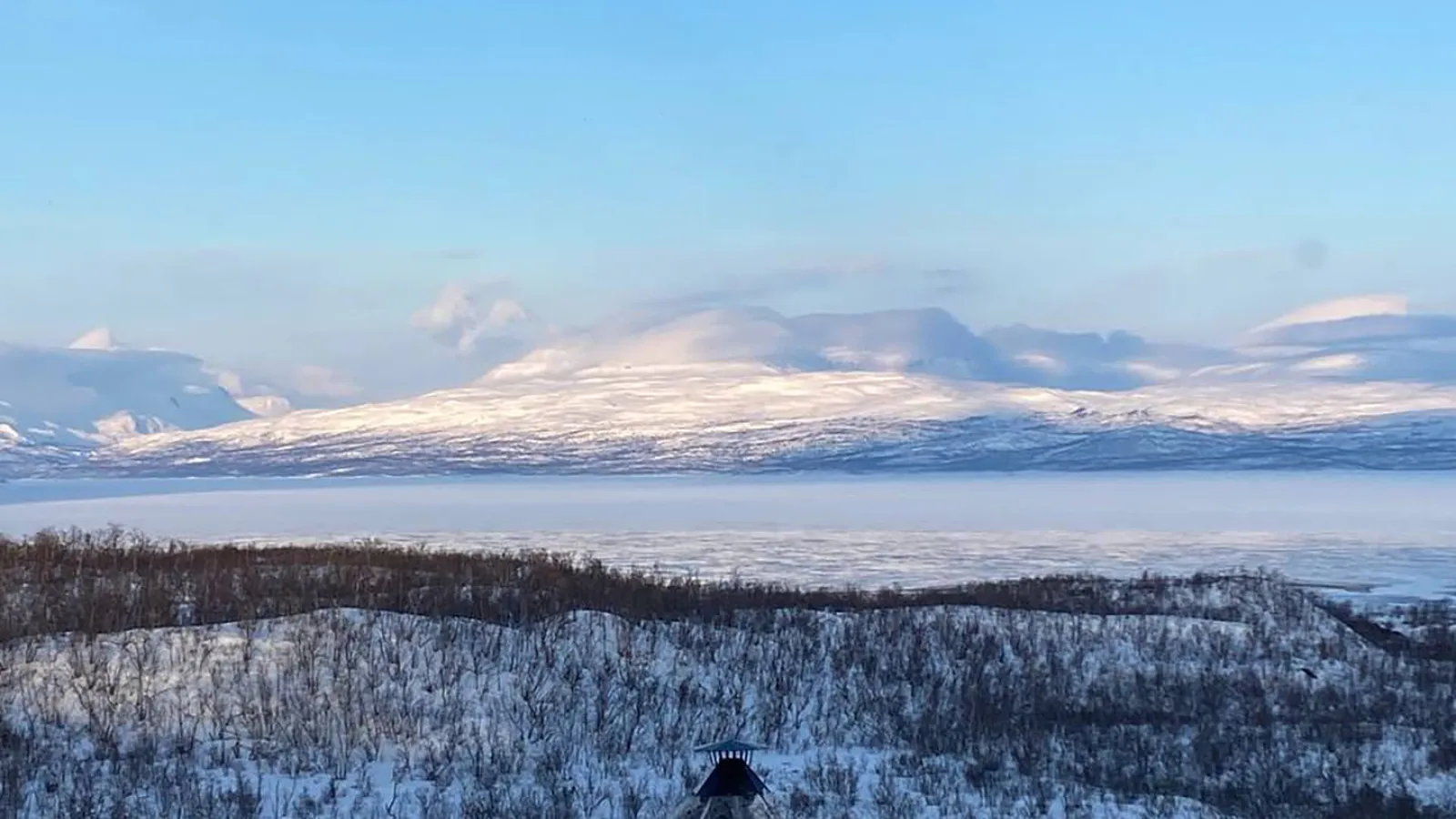 Eine kleine Holzhütte ist im Vordergrund einer verschneiten Landschaft. Kahle Bäume ragen aus dem Schnee heraus. Am Horizont sind Berge zu sehen die von Schnee bedeckt sind.