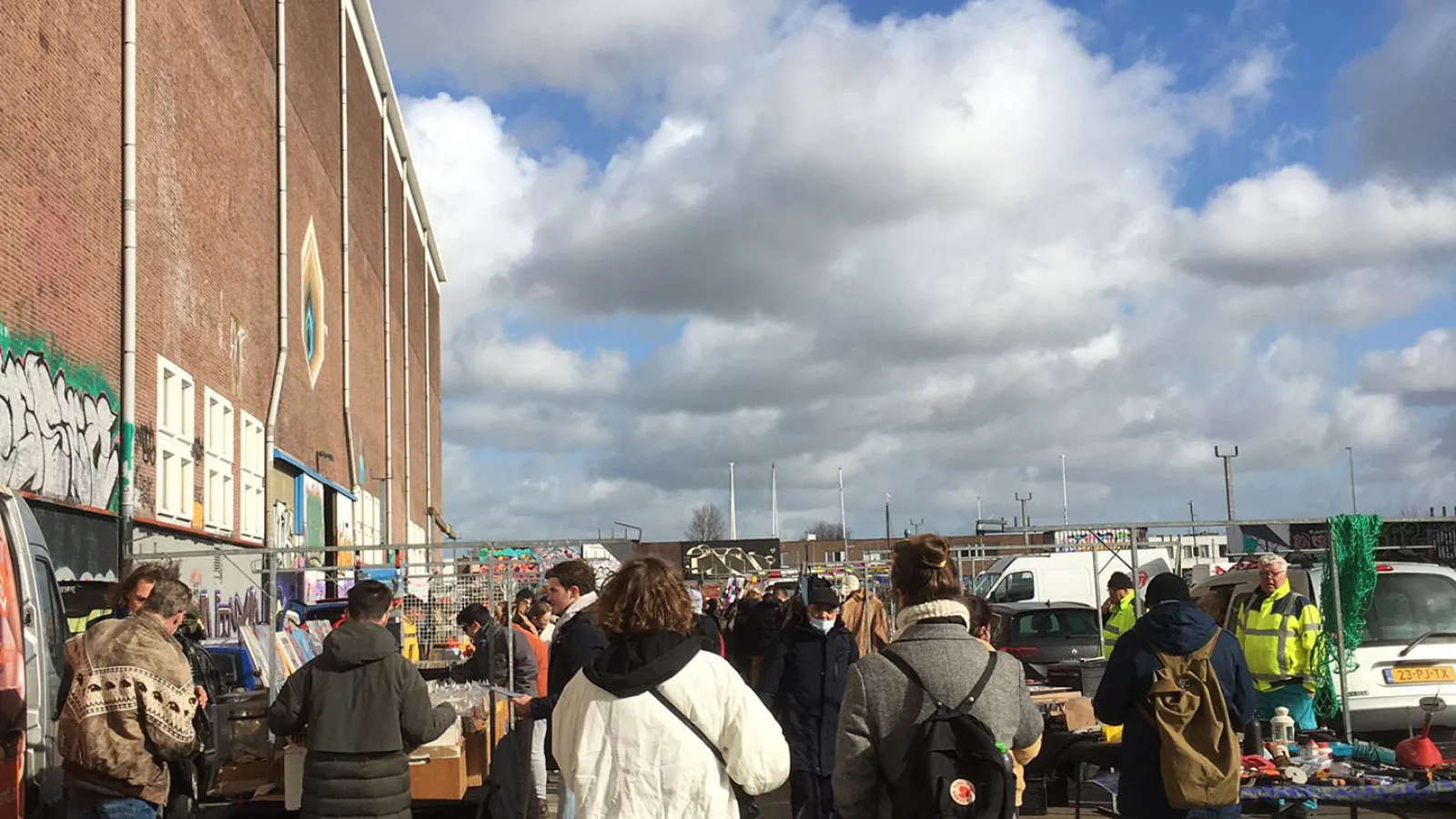 Im Vorderund sind zwei Menschen von hinten zu sehen die auf einen Flohmarkt zusteuern. Der Flohmarkt ist draußen. Der Himmel ist Blau und etwas bewölkt. 