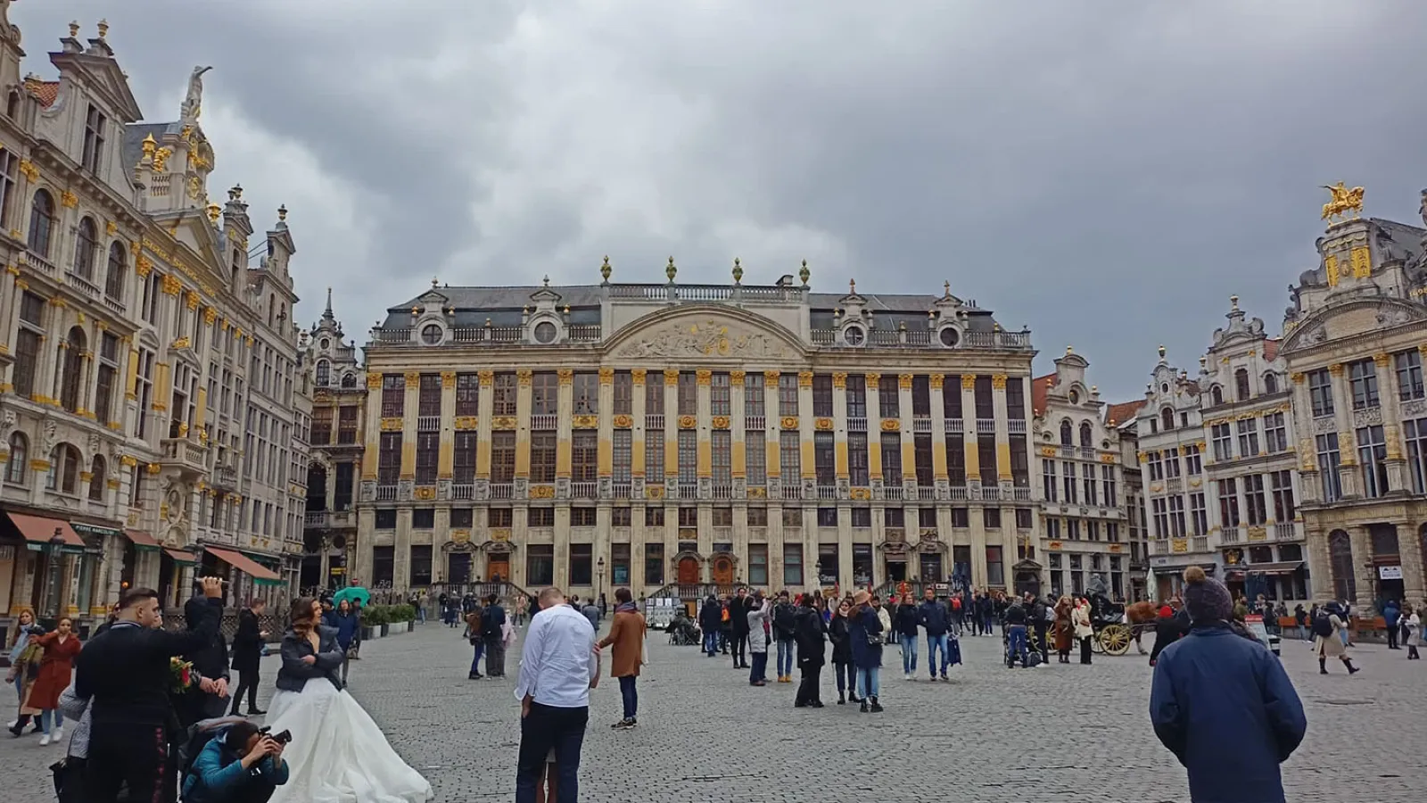 Ein Markplatz auf dem einige Menschen versammelt sind, u.a. im Vordergrund links eine Frau im Brautkleid. Der Blick ist aufd ein großes, altes, helles Gebäude gerichtet.