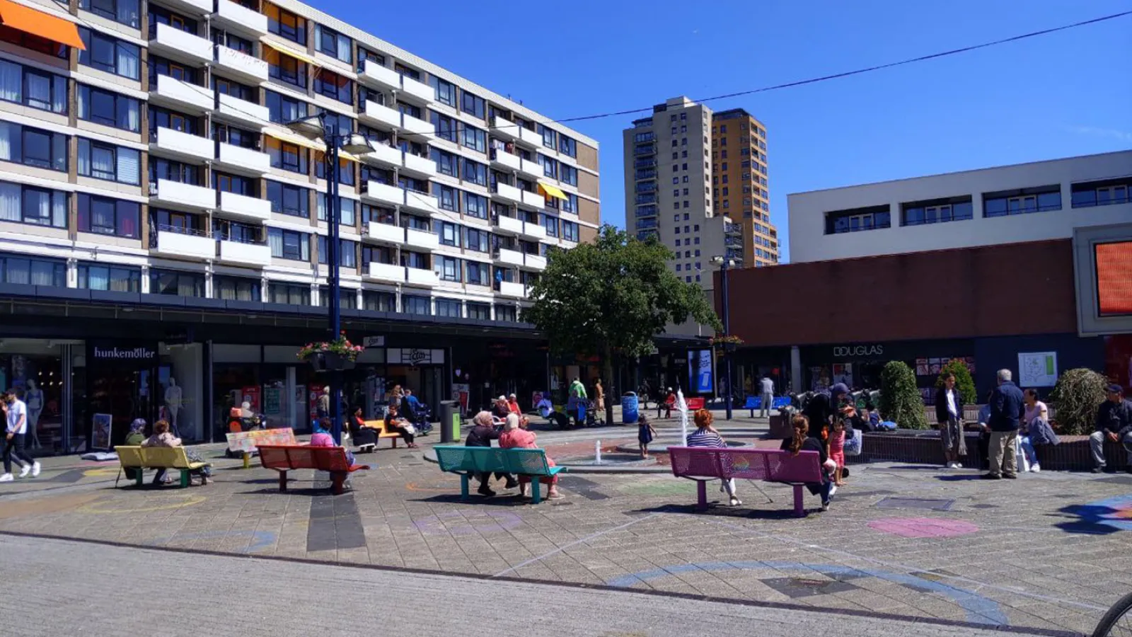 Auf einem Platz sind bunte Bänke. Auf einer Seite des Platzes sind weiße Häuser. Auf dem Platz sind nur vereinzelnd Menschen zu erkennen.