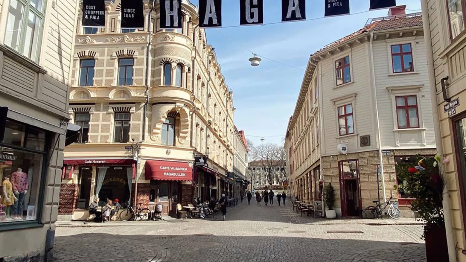 Eine Kopftseinpflasterstraße führt zu einer Kreuzung. Helle Gebäude stehen vor einem blauen Himmeln und werden von der Sonne beleuchtet. Über der Kreuzung sind Wimpeln gespannt auf denen die Buchstaben HAGA zu lesen sind.