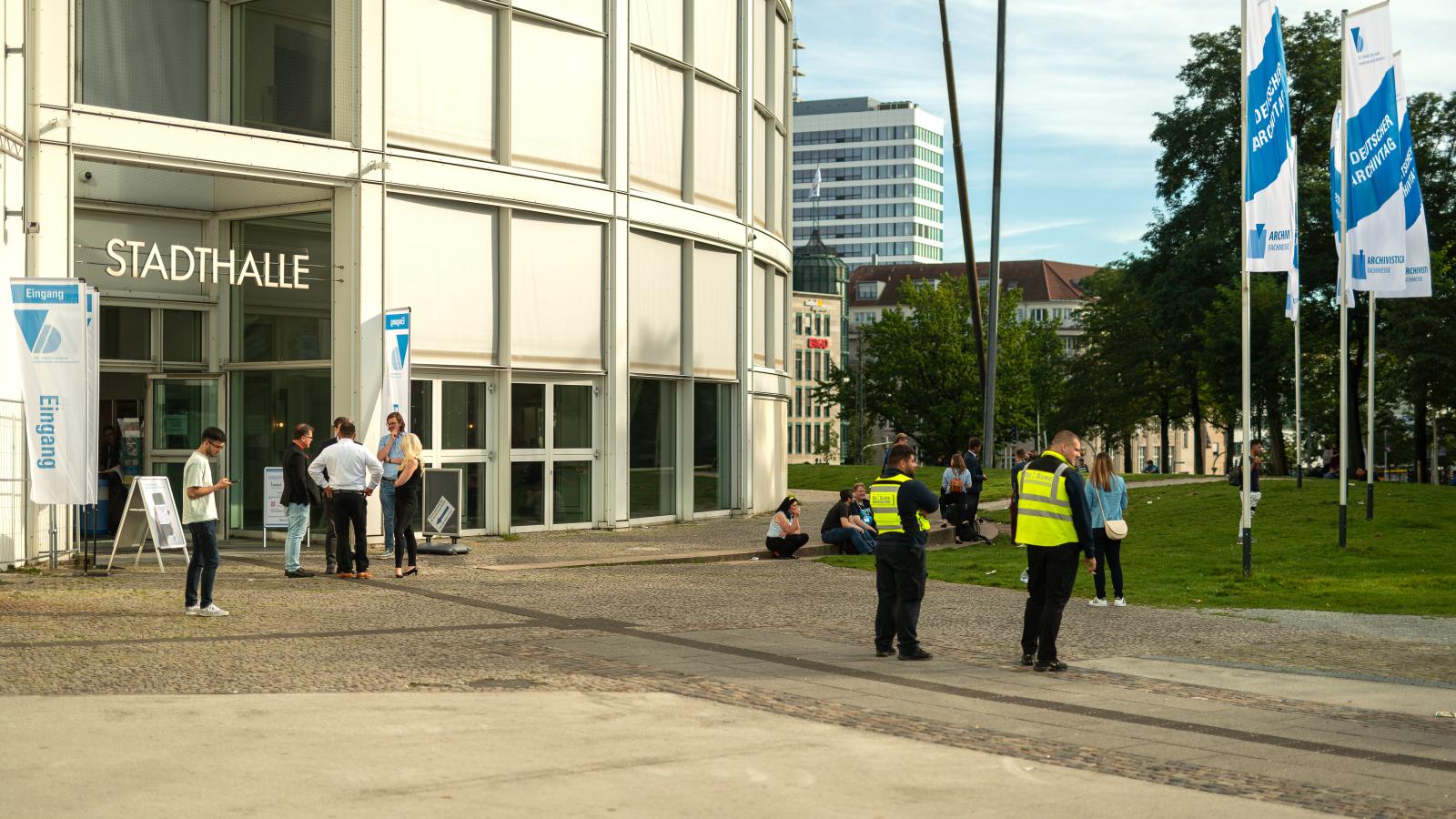 Ein Blick von Außen auf die Stadthalle Bielefeld, mit den Fahnen des deutschen Archivtags und Besuchenden vor dem Gebäude