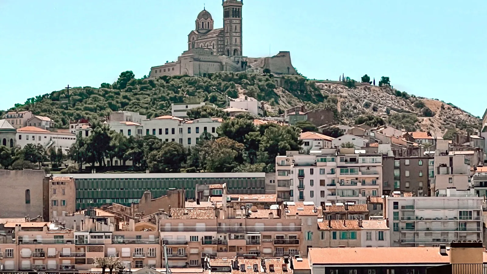 Der Blick führt vom unteren Rande de Bildes auf das klare Wasser und die Boote im Hafen und lenkt zu einer Häuserfront und letztendlich zu einem Berg Foch auf dem eine Kirche steht. Der Himmel ist klar und blau. 