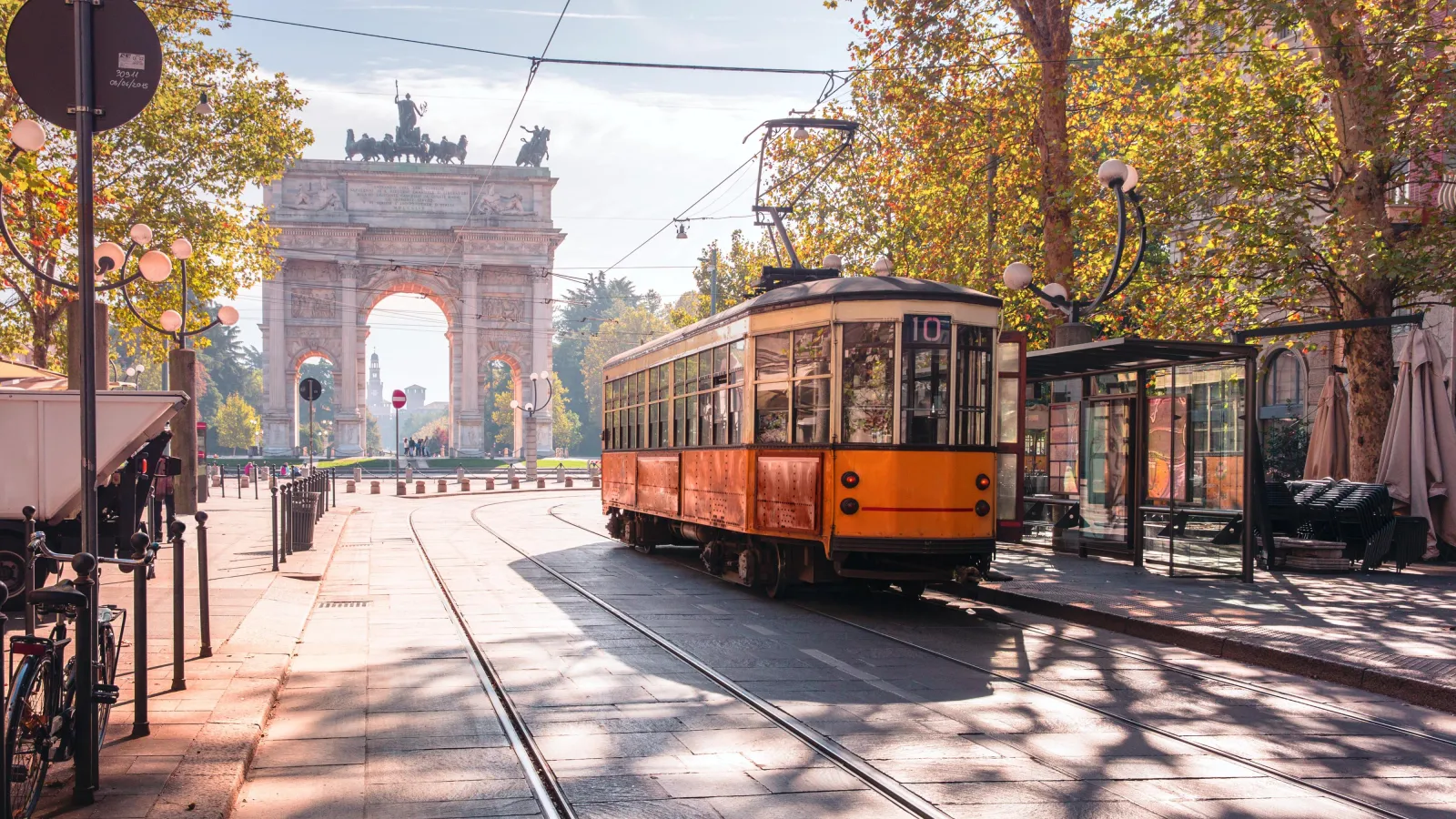 Eine alte rot und beige angemeldet Tram steht an der rechten Seite des Bildes vor einer Translation. Auf der linken Seite des Bildes ist das Tor "Arco della Pace" zu sehen. Es scheint ein herbstlicher Tag zu sein, die Sonne scheint und die Bäume haben braune Blätter. Die Straßen sind noch leer.