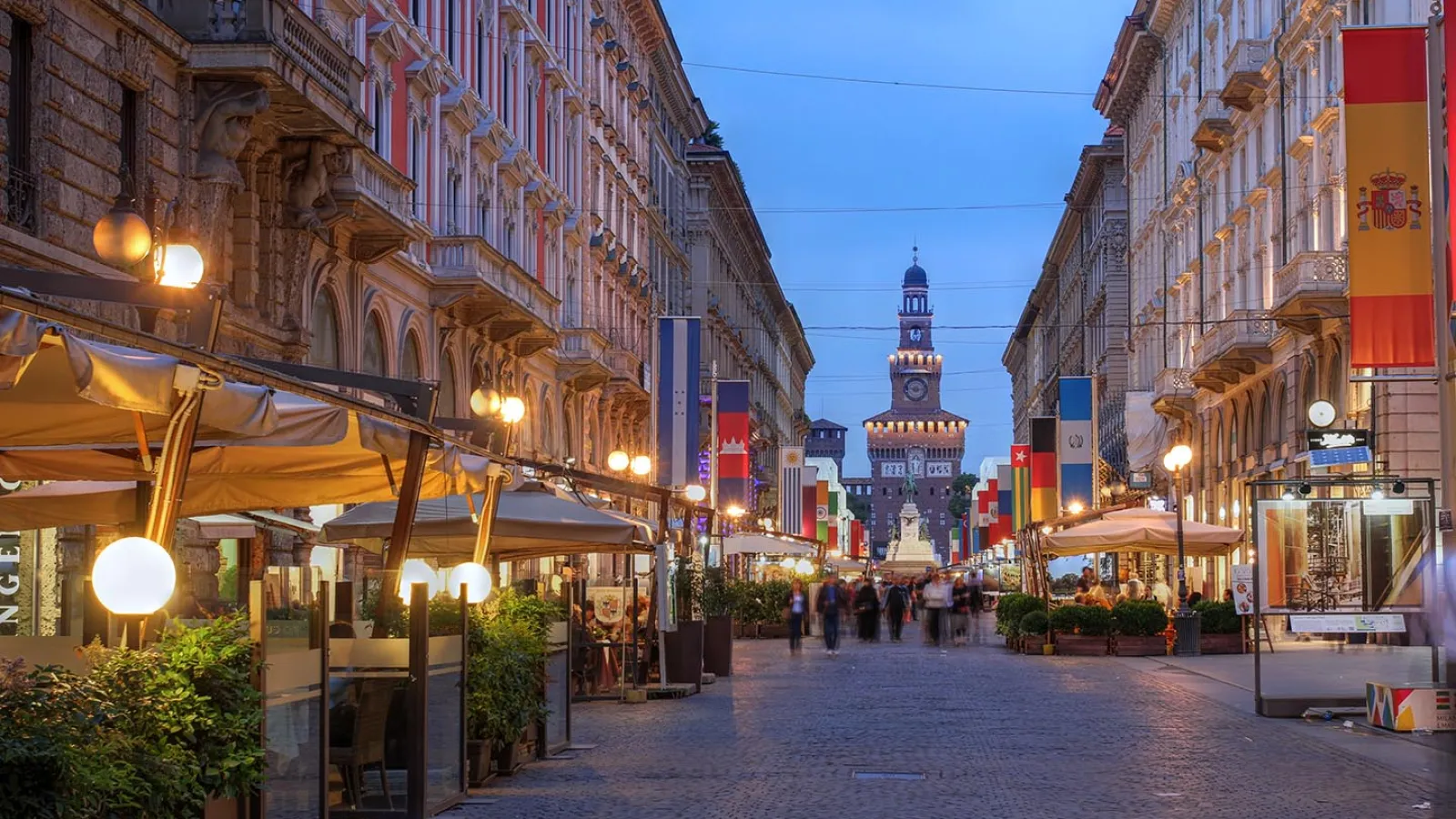 Der Blick führt eine Straße entlang. Es dämmert, die Lichter vor den Häusern sind an. Am Ende der Straße ist ein Turm aus Stein zu sehen.