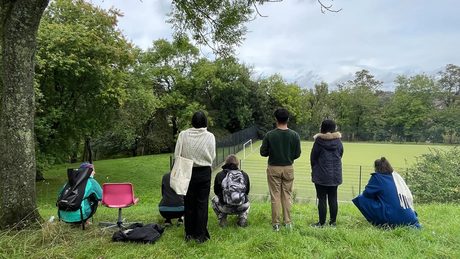 Eine Gruppe Menschen ist am Rande eines grünen Hügels positioniert. Einige stehen, einige sind in der Hocke. Alle gucken von der Kamera weg: sie betrachten den Blick. Zwischen den Menschen ist ein knall pinker Stuhl mit aufgereiht. 