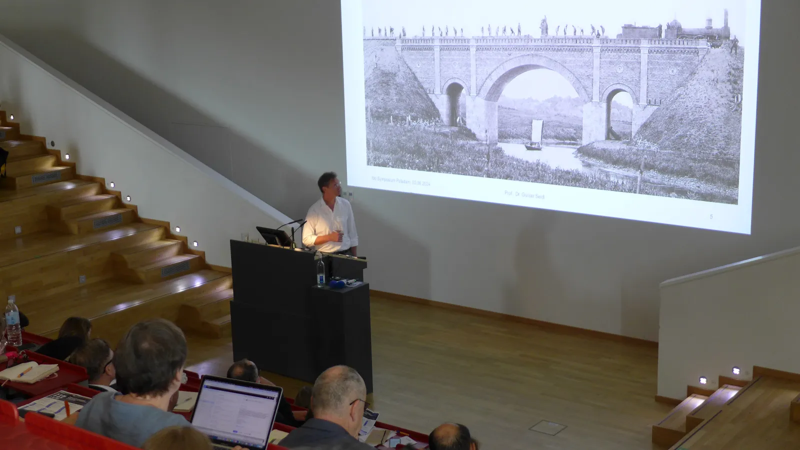 Herr Prof. Dr. Günter Seidl hält einen Vortrag im Hörsaal, an die Wand wird ein Bild projiziert. Zuhörerende schauen sich den Vortrag an