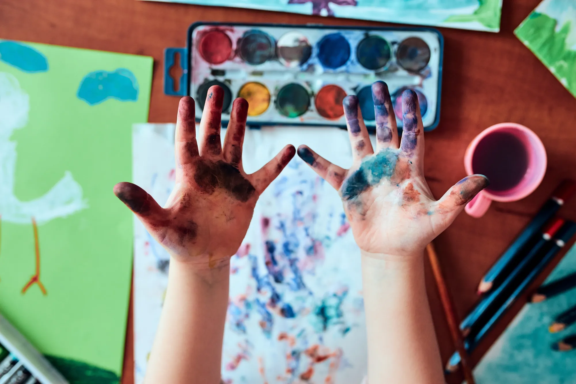 zwei bemalte Kinderhände vor Tischplatte mit Aquarellfarben, Malpapier und Pinseln