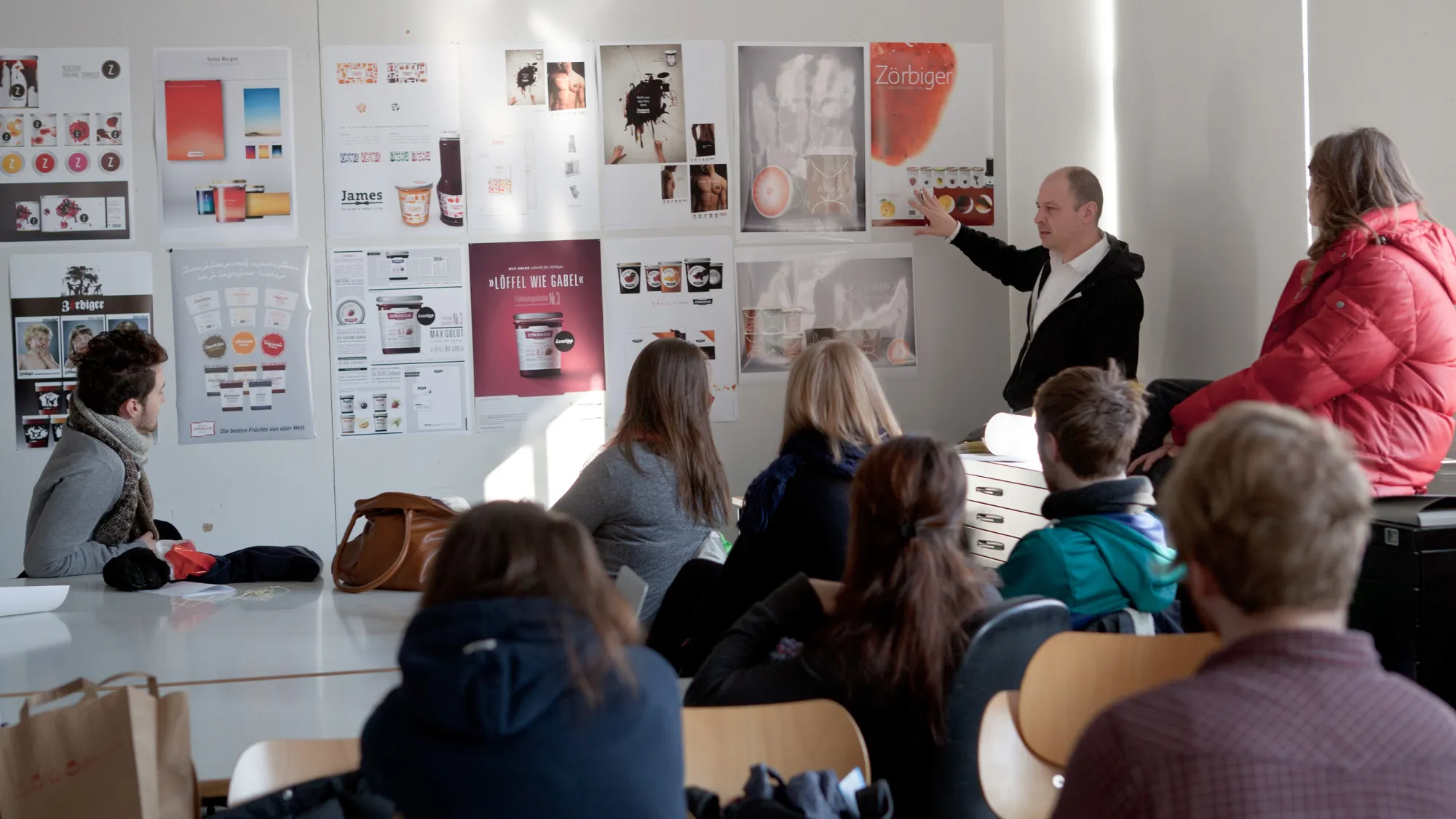 Lehrender zeigt vor Studierenden auf Entwürfe an einer Wand aus dem Studiengang Kommunikationsdesign