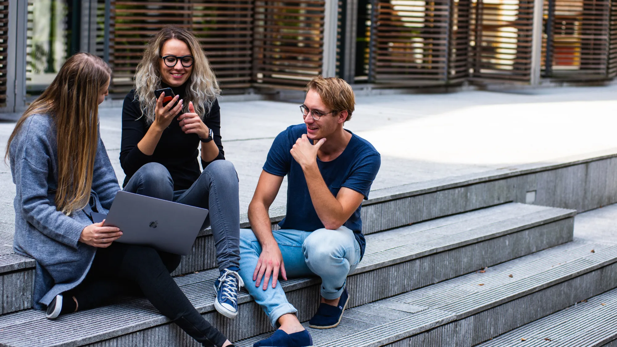 Drei Personen sitzen in einer Gruppe auf einer Treppe; eine Person hat einen Laptop auf dem Schoß