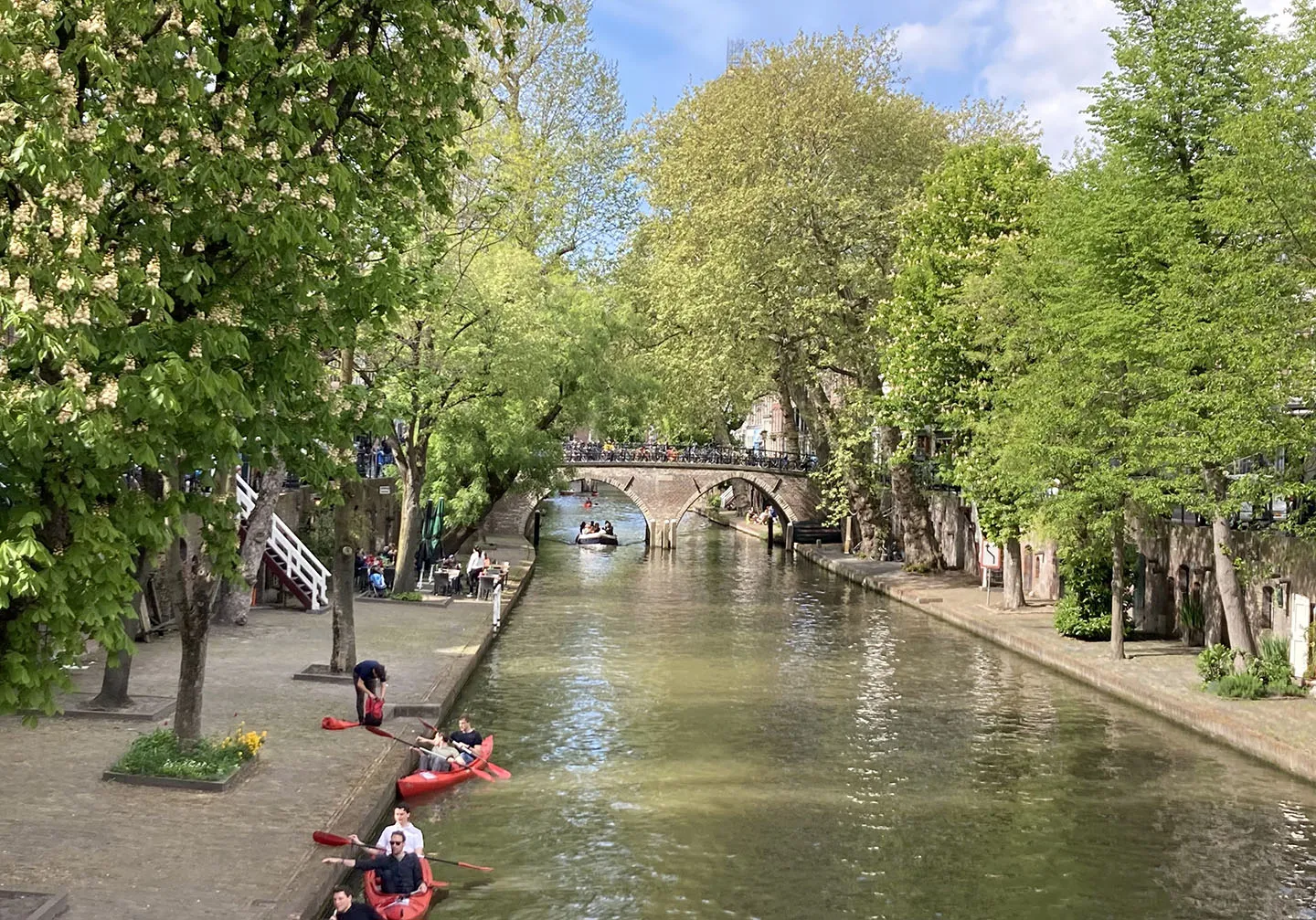 Man blickt auf einen stillen Kanal und eine kleine Brücke aus Stein die diesen überquert. Das Ufer ist mit Bäumen bepflanzt die viele Grüne Blätter tragen. Zwei Personen paddeln einzeln im Kanu.