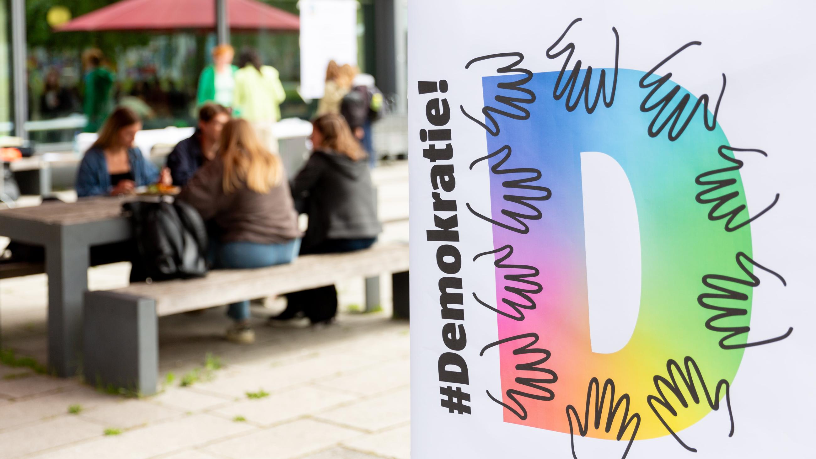 Personen sitzten auf einer Bank vor der Mensa, davor ein Plakat mit dem Titel "Hashtag Demokratie"