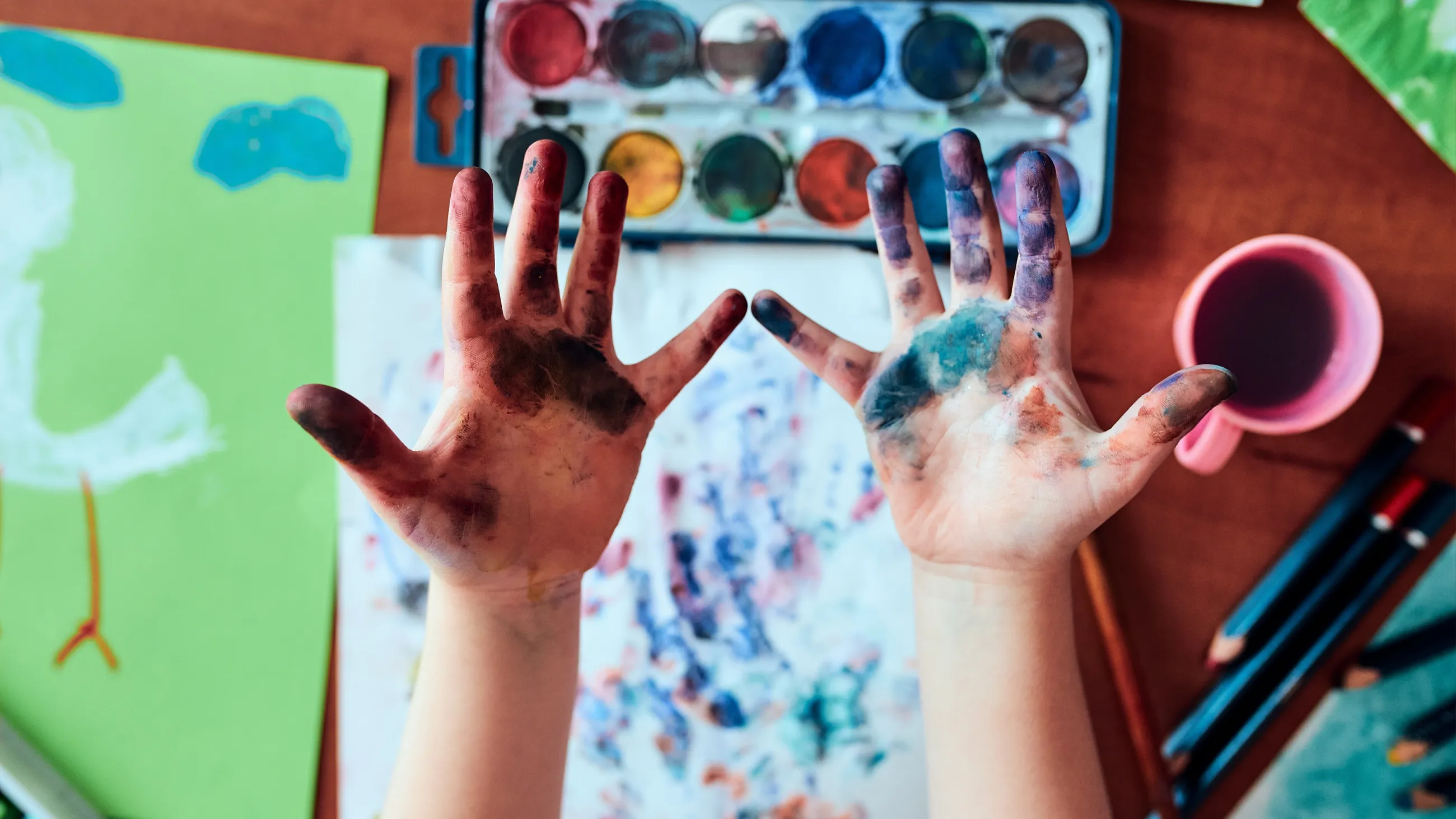 zwei bemalte Kinderhände vor Tischplatte mit Aquarellfarben, Malpapier und Pinseln