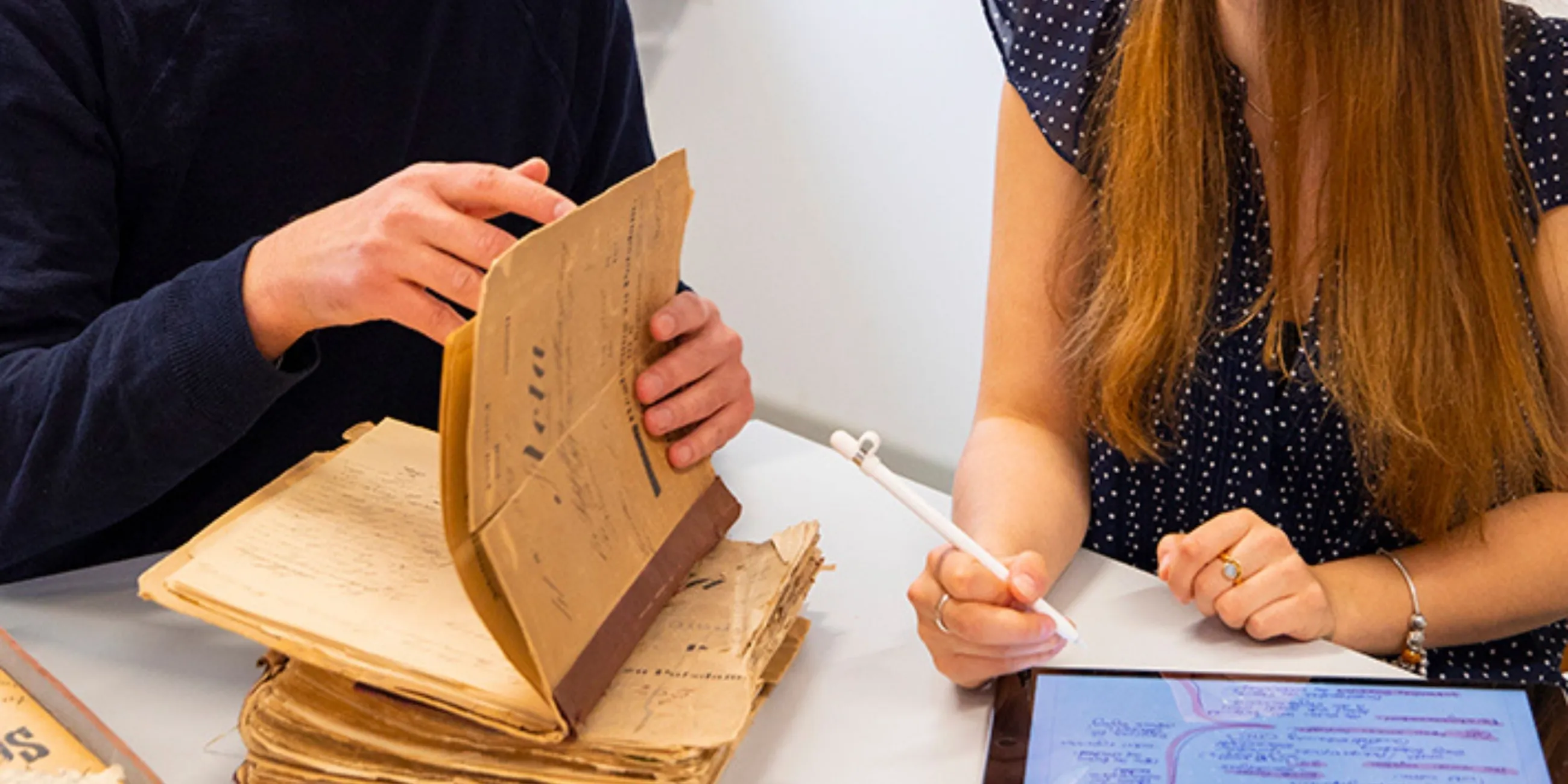 Zwei Studierende arbeiten mit historischen Dokumenten. Eine Person davon blättert um. Eine weitere gibt Daten in ein Tablet ein. 