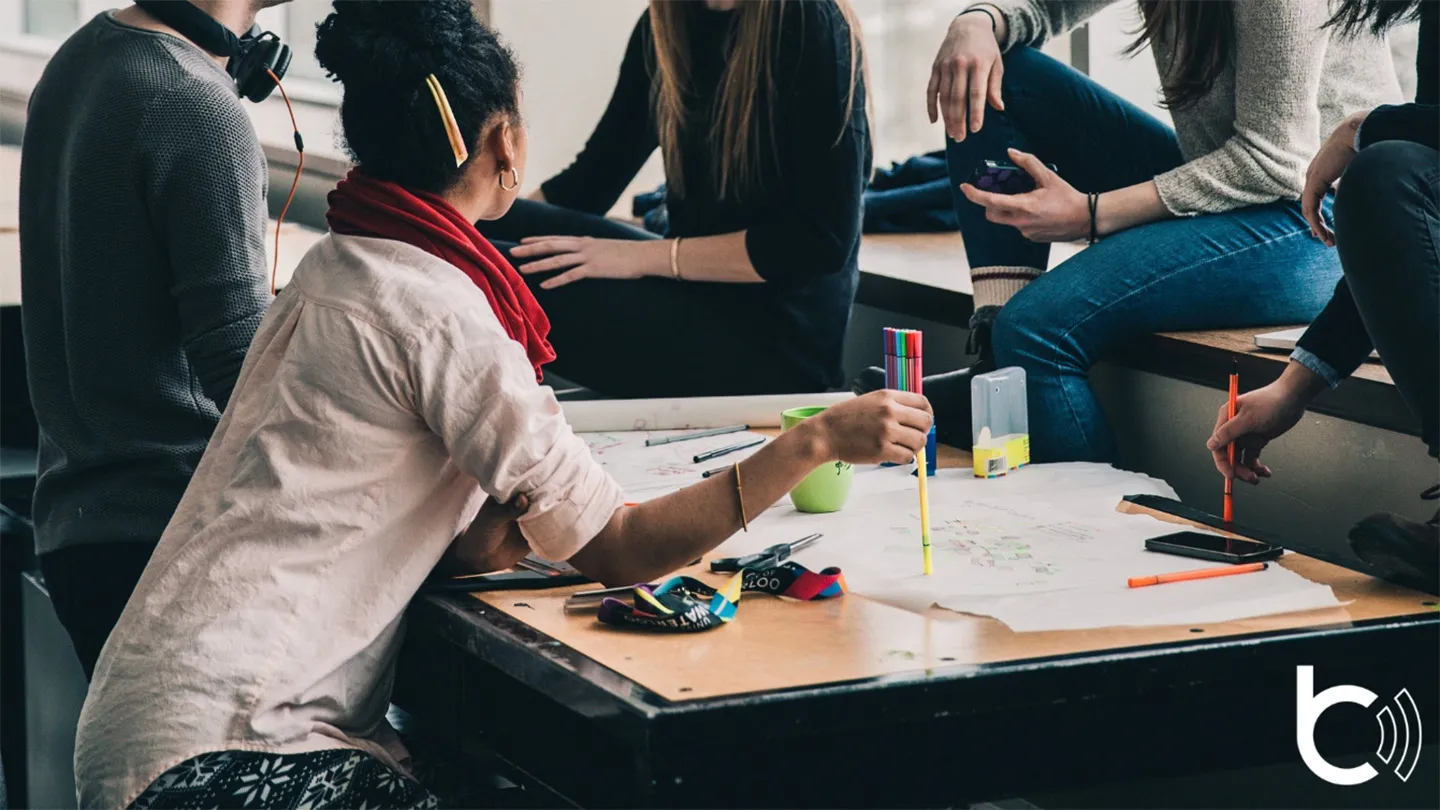 Fünf Personen sitzen um einen Tisch herum; auf dem Tisch sind beschriftete Papiere und bunte Stifte zu sehen